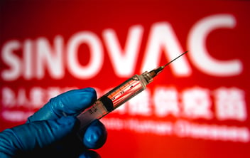 A gloved hand holds a vaccine syringe, in front of a large red sign that says 
